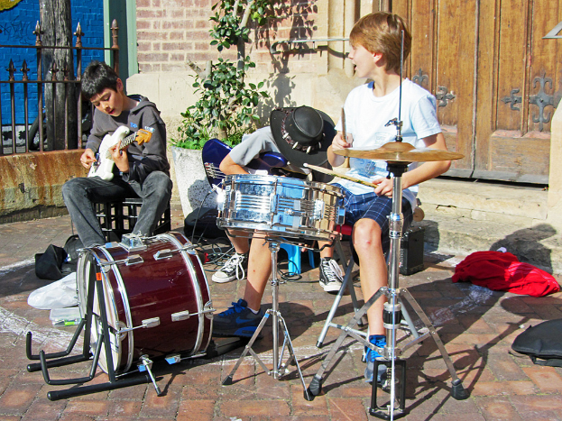 Drei Jungen auf Stühlen sitzend und Musikinstrumente spielend vor einer geschlossenen Tür mit einem Blumentopf in der Nähe.