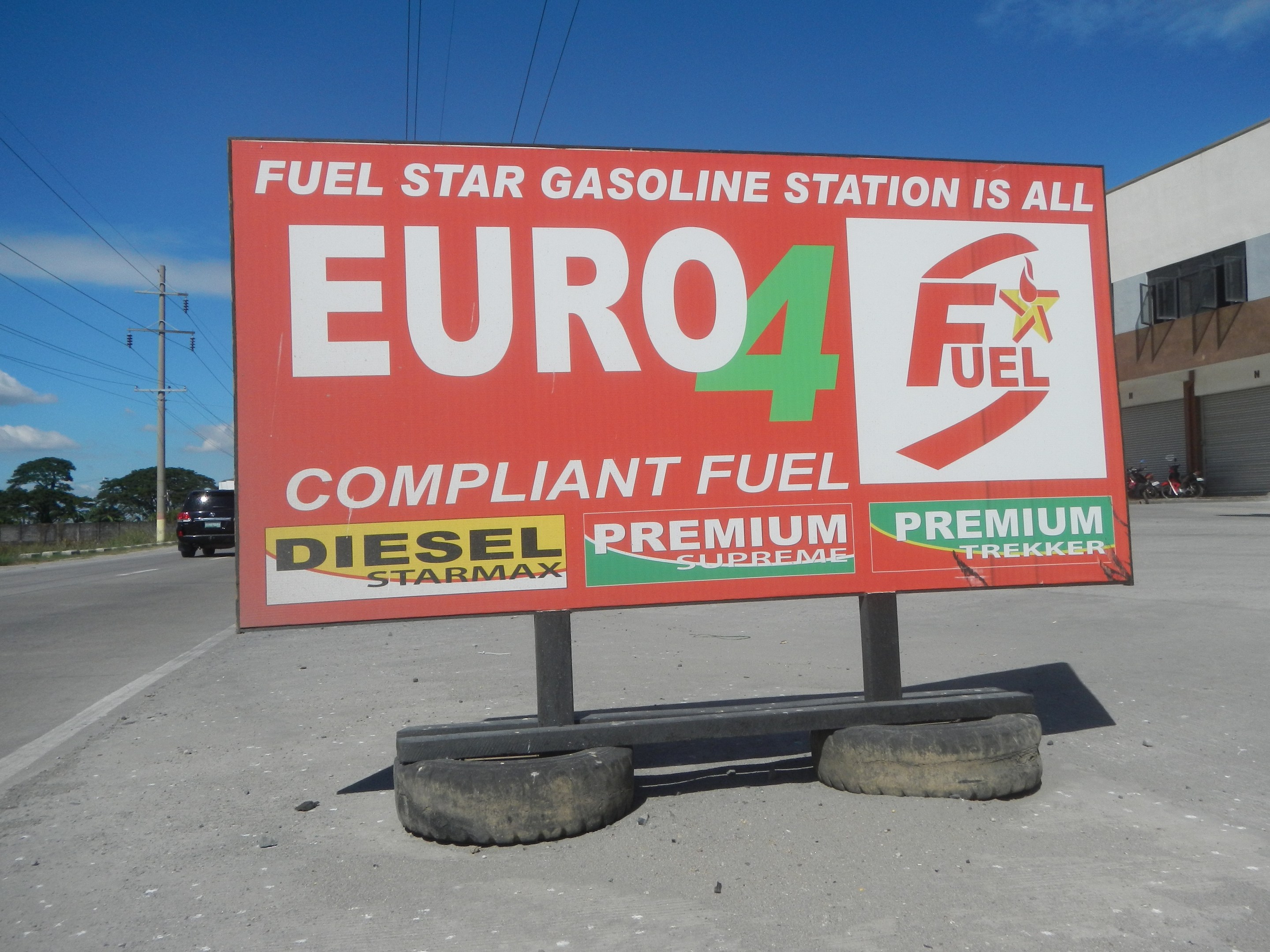 Tankstelle mit "Euro4 Fuel"-Schild, Fahrzeuge auf der Straße, Strommasten, Bäume, ein Gebäude und Himmel im Hintergrund.