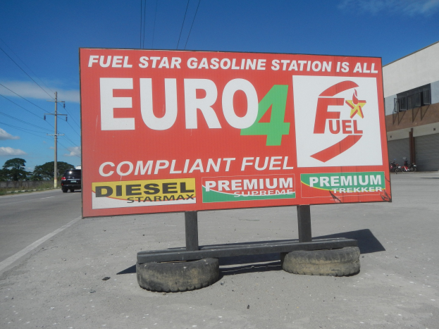 Tankstelle mit "Euro4 Fuel"-Schild, Fahrzeuge auf der Straße, Strommasten, Bäume, ein Gebäude und Himmel im Hintergrund.