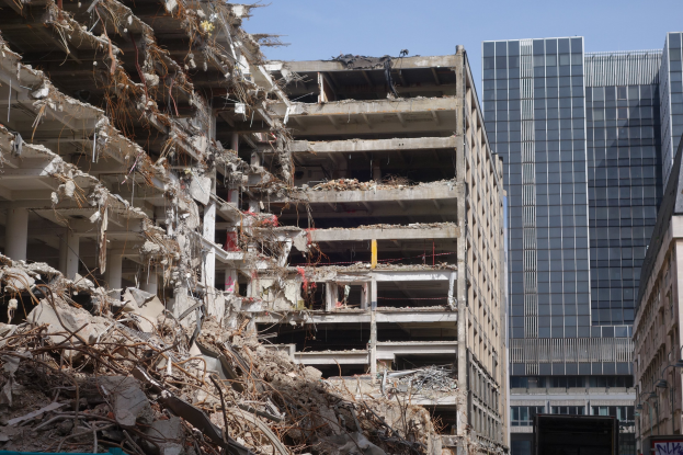 Ein großes Gebäude wird inmitten einer Stadt abgerissen, umgeben von intakten Gebäuden, Straßeninfrastruktur, Fahrzeugen, Bäumen und einem klaren Himmel.