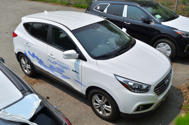 Three cars parked side by side in a lot, with a mesh fence and grass in the background, one displaying hydrogen-powered vehicle text.