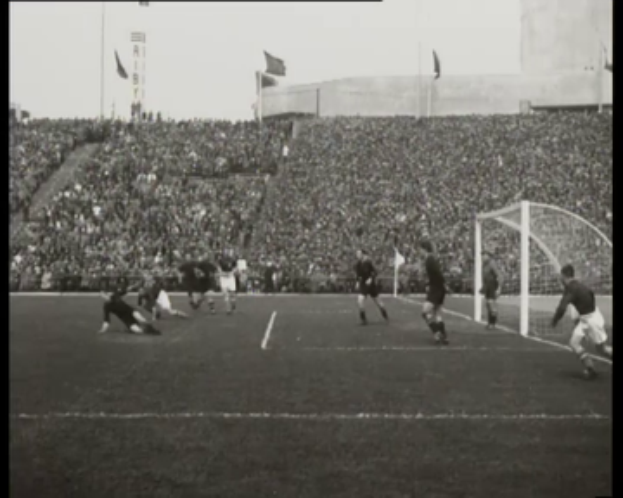 Ein Schwarz-Weiß-Foto eines WM-Finalspiels 1958 zwischen Manchester United und Liverpool mit Spielern auf dem Feld, einem Torpfosten rechts, Zuschauern in den Rängen und Fahnen im Hintergrund.