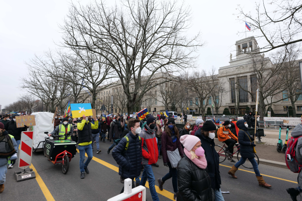 Eine große Gruppe von Menschen nimmt an einer Protestdemo auf einer Straße in Washington, D.C. teil, einige halten Schilder und Banner, andere fahren Fahrräder, und es sind Schilder, Bäume und ein klarer blauer Himmel im Hintergrund zu sehen.