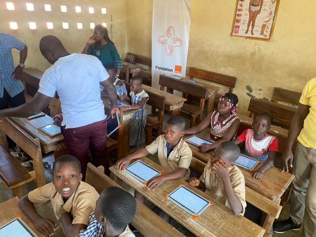 Eine Gruppe von Kindern sitzt an Tischen in einem Klassenzimmer und nutzt Tablets, während einige Menschen um sie herumstehen und ein Banner und ein Plakat im Hintergrund zu sehen sind.
