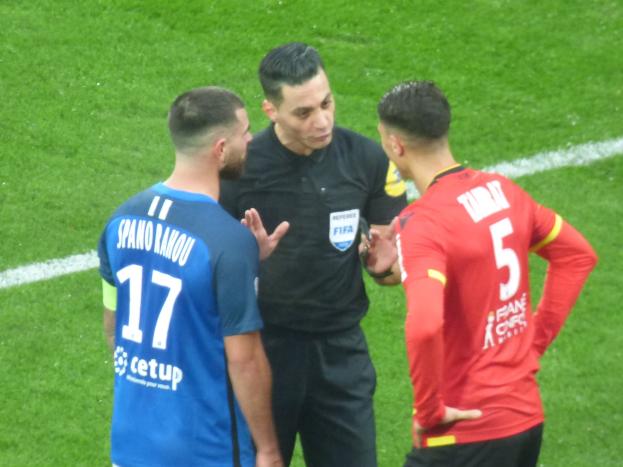 Schiedsrichter spricht mit zwei Männern auf einem Fußballfeld, einer trägt ein schwarzes Hemd und Hose, der andere ein blau-rotes T-Shirt mit Text und Brille.