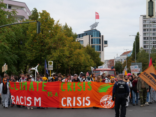 Gruppe von Menschen marschiert auf einer von Bäumen gesäumten Straße und hält ein 'Klimakrise ist eine Krise'-Schild hoch, mit parkenden Fahrzeugen, Gebäuden und einem klaren blauen Himmel im Hintergrund.