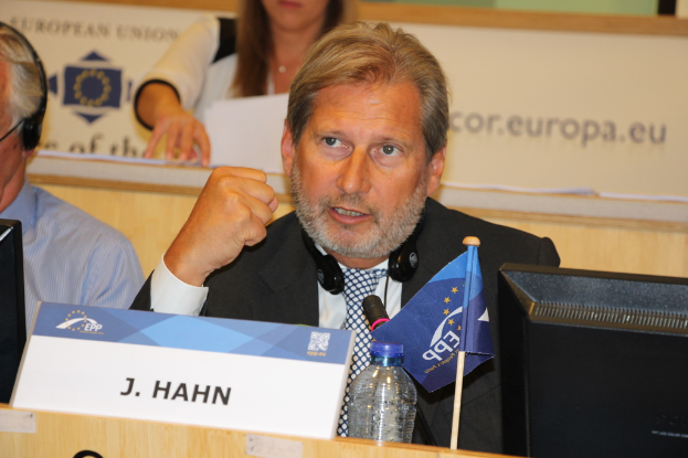 Ein Mann in einem schwarzen Blazer, weißem Hemd und schwarzer und weißer Krawatte spricht in ein Mikrofon, trägt einen Headset um den Hals und hat vor sich auf einem Tisch eine Wasserflasche, eine Flagge, einen Laptop und ein Namensschild; links neben ihm arbeitet ein Mann in einem blauen Hemd an einem System und trägt Kopfhörer; hinter ihnen hält eine Frau Papiere.