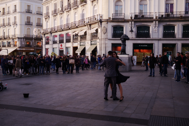 Ein Mann und eine Frau tanzen auf einem belebten Stadtplatz, umgeben von Zuschauern, mit einem Kinderwagen, Eimer und Stangen links und Gebäuden, Schildern, einer Statue und einem bewölktem Himmel im Hintergrund.