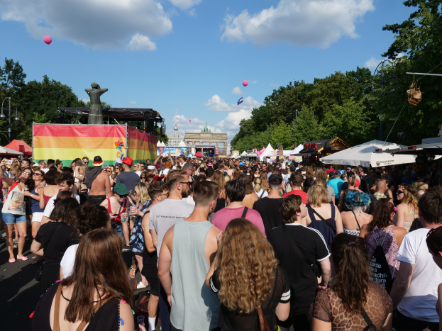 Eine große Menschenmenge, die eine Straße mit Zelten, Bäumen, Pfählen, Lichtern und einer Statue säumt, mit Gebäuden im Hintergrund und einem bewölkten Himmel mit Luftballons während des Christopher Street Day in Berlin.