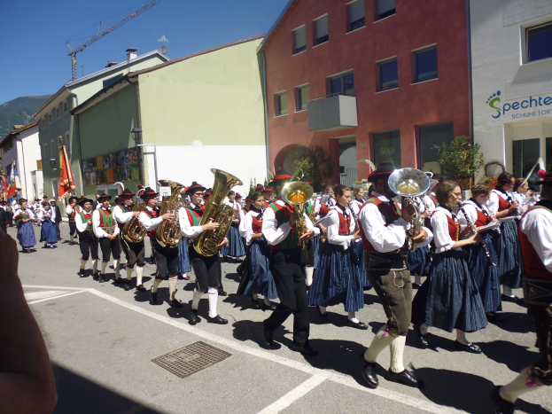 Eine Gruppe von Menschen in traditioneller bayrischer Tracht, die auf der Straße Musikinstrumente spielen und dabei durch eine Straße mit Gebäuden gehen, einige halten Fahnen, mit einem Hügel und einem klaren blauen Himmel im Hintergrund.