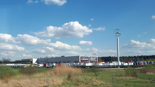 Ein Mercedes-Benz-Werk in Deutschland mit Fahrzeugen auf der Straße, umgeben von Gras, Pflanzen, Bäumen, Gebäuden und Polen, einschließlich eines mit einem Logo, unter einem bewölkten Himmel.