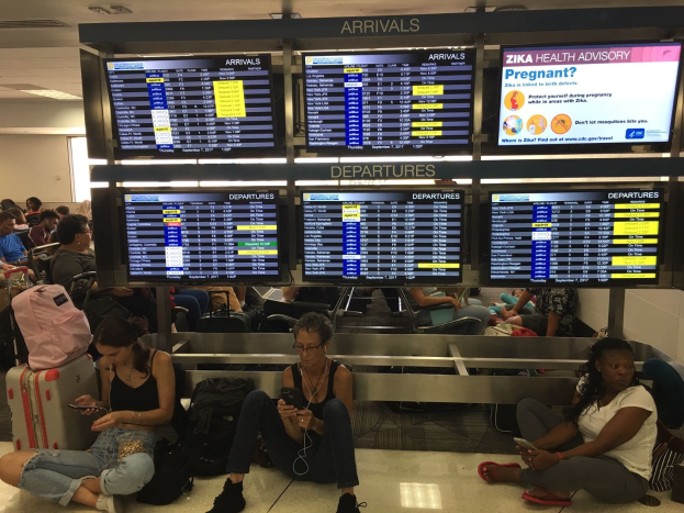 Gruppe von Menschen auf einem Flughafenboden sitzend, einige halten Handys, mit verstreuten Taschen und Zika-Virus-Informationen auf Bildschirmen im Hintergrund.