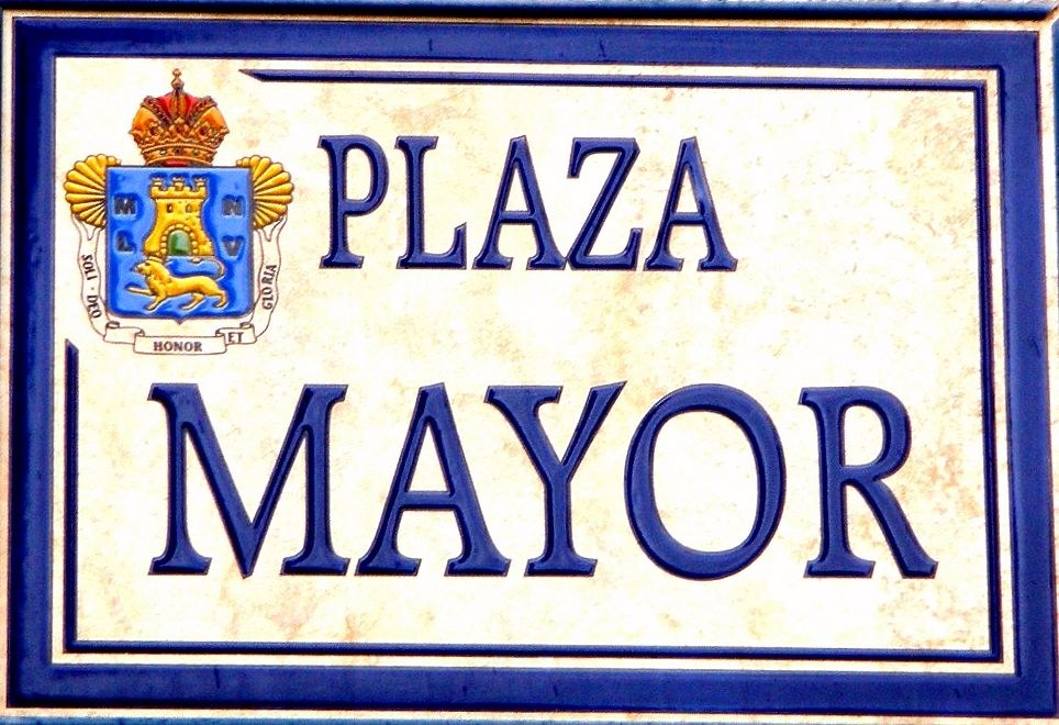 Plakette an einem Gebäude mit der Aufschrift "Plaza Mayor" mit einem blauen Rahmen, weißem Hintergrund, schwarzer fettgedruckter Schrift und einem gekrönten Wappen-Logo.