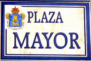Plakette an einem Gebäude mit der Aufschrift "Plaza Mayor" mit einem blauen Rahmen, weißem Hintergrund, schwarzer fettgedruckter Schrift und einem gekrönten Wappen-Logo.
