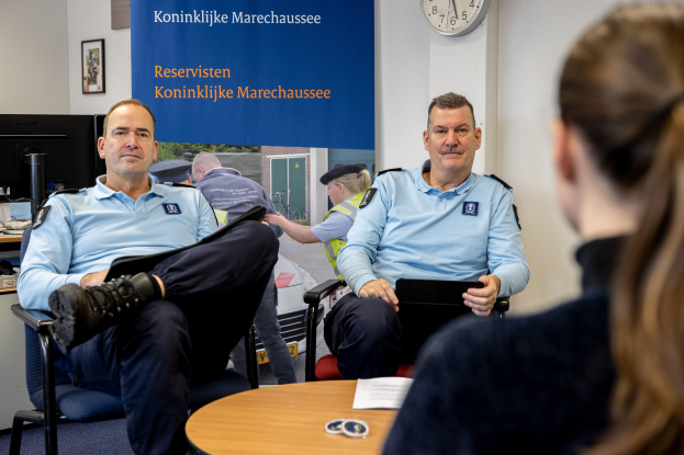 Zwei Polizeibeamte sitzen an einem Tisch mit Papieren und Gegenständen und sprechen mit einer Frau, mit einer Fahne, Uhr und Fotorahmen an der Wand und einem weiteren Tisch mit einem Monitor und anderen Gegenständen links daneben.