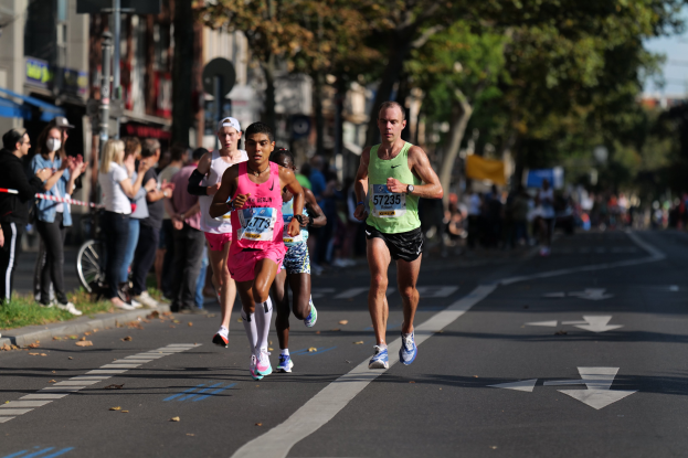 Gruppe von Läufern bei einem Marathon auf einer Stadtstraße mit Zuschauern, Bäumen, Gebäuden und einem Fahrrad im Hintergrund.