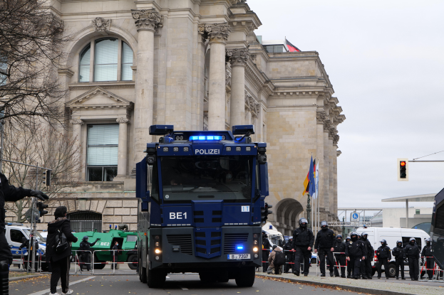 Eine Gruppe von Polizisten steht vor einem großen Gebäude mit Fenstern, Säulen und Bögen, mit Fahrzeugen auf der Straße und einer Person mit einer Kamera auf der linken Seite, unter einem klaren blauen Himmel mit Bäumen, Verkehrsampeln und Fahnen im Hintergrund.