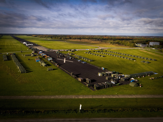 Luftaufnahme einer Militärbasis in den Niederlanden mit Fahrzeugen auf Straßen, Grasflächen, Bäumen, Gebäuden und einer bewölkten Himmel.