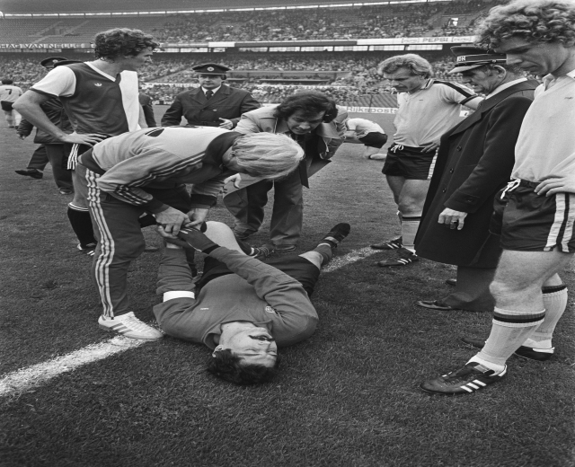 Schwarzes und weißes Bild eines Fußballspielers, der auf dem Boden liegt und von einer Gruppe Männer und einem Stadionpublikum umgeben ist, der scheinbar verletzt ist.