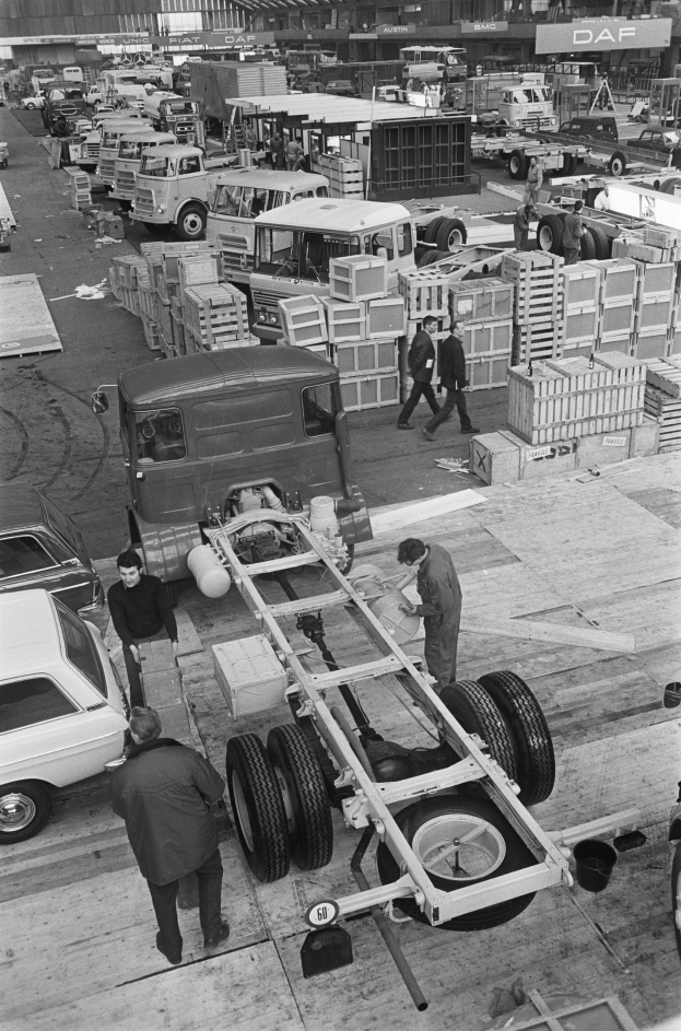 Schwarze und weiße Fotografie von Männern, die an einem Lkw in einer Fabrik arbeiten, mit Fahrzeugen und Menschen im Hintergrund.