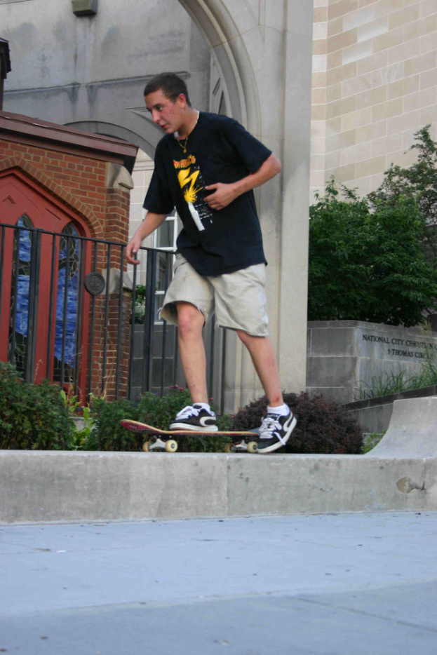 Ein Mann fährt mit dem Skateboard vertikal an einer Wand hoch mit einer Plattform unten, mit einem Haus, Grill, Pflanzen, Bäumen, einer Backsteinwand und einer Säule im Hintergrund.