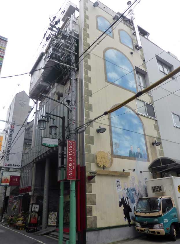 Ein Lastwagen parkt an der Straße neben einem hohen Gebäude mit einer Wandmalerei, mit Strommasten, Kabeln, Straßeleuchten, Namensschildern, Kraftfahrzeugen, Topfpflanzen und einem klaren blauen Himmel im Hintergrund.