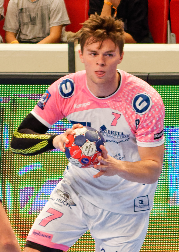 Ein Mann in einem rosa Hemd hält einen Fußball, mit der Hand eines anderen Menschen auf der linken Seite und einigen Menschen im Hintergrund, die auf Stühlen sitzen.