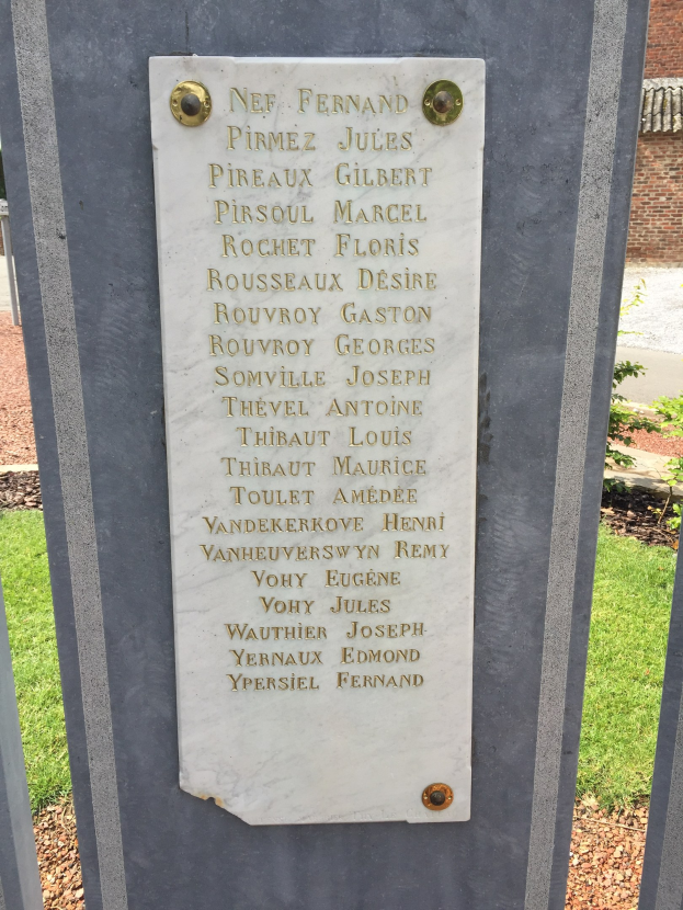 Steinbank mit einer Gedenktafel vor einem Backsteingebäude, umgeben von Grün.