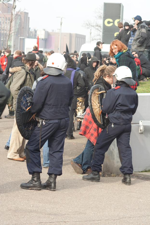 Eine Gruppe von Menschen, die auf einer Straße gehen, mit zwei Personen in der Front, die wie Polizisten aussehen, Gebäuden im Hintergrund und Boden unten.