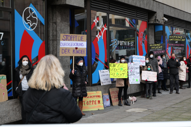 Maskierte Menschen mit Schildern und Plakaten protestieren vor einem Glasgebäude, mit Kameras und einem Hund anwesend, wahrscheinlich gegen eine deutsche Schulmaskenpflicht.
