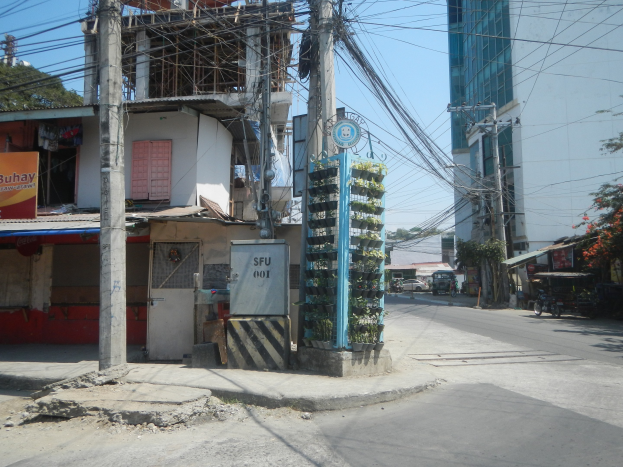 Eine Stadtstraße mit Gebäuden, Strommasten, Kabeln, Namensschildern, Fahrzeugen, Bäumen und einem Himmel als Hintergrund, mit einem Gebäude mit Drähten an der Seite im Vordergrund.