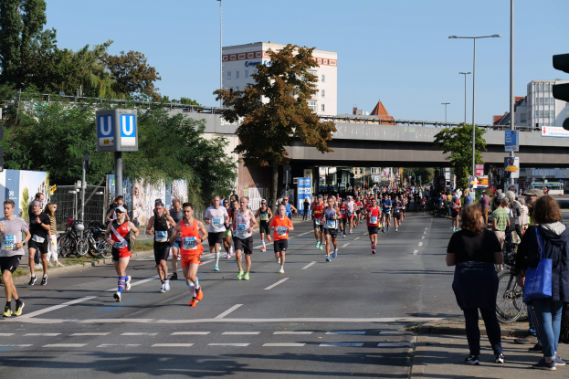 Gruppe von Menschen, die auf einer von Bäumen gesäumten Straße mit Laternen, Schildern, Fahrrädern, einem Zaun, Gras, einer Brücke, Gebäuden und einem klaren blauen Himmel laufen.