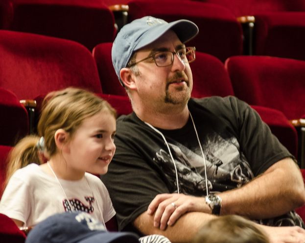 Ein Mann mit Brille, Uhr und Mütze sitzt mit einem strahlenden kleinen Mädchen in einem Kinosaal, mit Stühlen und anderen Menschen im Hintergrund.