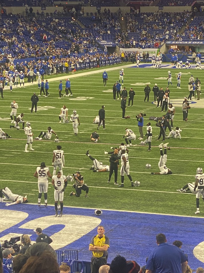 Ein Fußballspiel auf einem Stadionfeld mit einigen stehenden und sitzenden Menschen im Hintergrund, während eine Person eine Kamera für die Live-Übertragung hält.