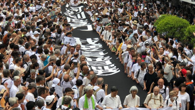 Eine große Gruppe von Menschen marschiert auf der Straße, hält Protestschilder und Banner in der Hand, mit Grünpflanzen auf der rechten Seite und einem Gebäude im Hintergrund.