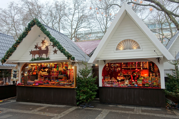Ein kleines weißes Gebäude mit festlicher Dekoration und Lichtern im Inneren, umgeben von Häusern, Bäumen und einem klaren blauen Himmel, mit einem Weihnachtsmarkt davor.