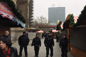 Eine Gruppe von Polizisten in schwarzen Uniformen und Mützen steht vor einem geschmützten Weihnachtsmarkt in Berlin, Deutschland, mit festlichen Dekoelementen, Gebäuden, Bäumen und einem klaren blauen Himmel im Hintergrund; ein Schild mit Text ist links sichtbar.