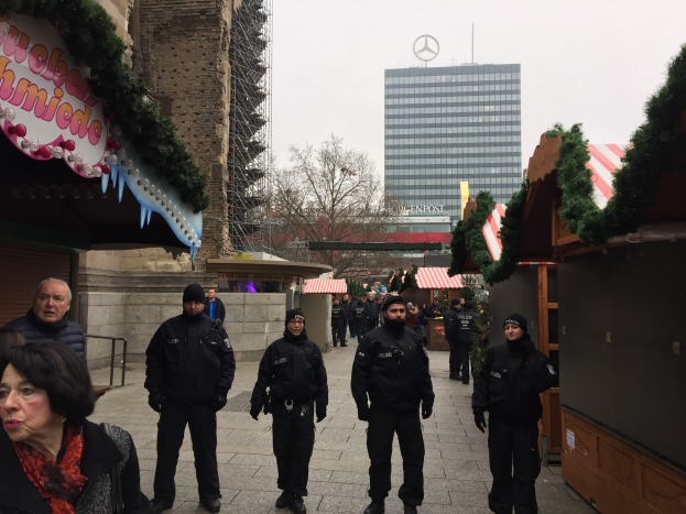 Eine Gruppe von Polizisten in schwarzen Uniformen und Mützen steht vor einem geschmützten Weihnachtsmarkt in Berlin, Deutschland, mit festlichen Dekoelementen, Gebäuden, Bäumen und einem klaren blauen Himmel im Hintergrund; ein Schild mit Text ist links sichtbar.