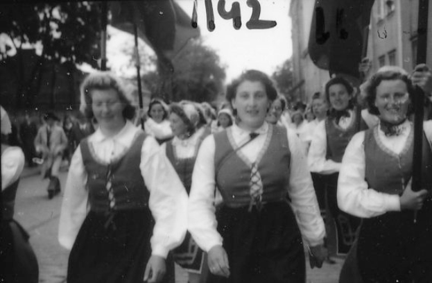 Eine Gruppe von Frauen in traditioneller bayrischer Tracht geht eine Straße entlang, lächelt und ist von Bäumen und Gebäuden umgeben, auf einem Schwarz-Weiß-Bild.