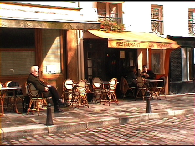 Ein Mann sitzt auf einem Stuhl auf einem Gehweg mit Tischen und Stühlen vor einem Gebäude mit einer Tafel, auf der "Restaurant" steht.