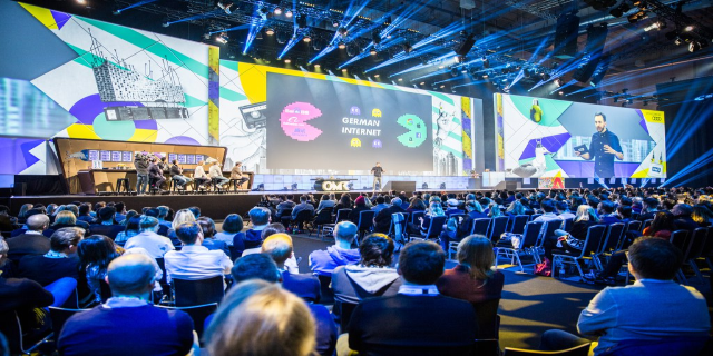 Eine große Menschenmenge, die auf Stühlen vor einer Bühne sitzt, mit einigen Personen, die auf dem Podium in der Vordergrund stehen, einem Tisch mit verschiedenen Gegenständen links und Bildschirmen und Lichtern im Hintergrund auf der Deutschen Internet-Konferenz 2019.