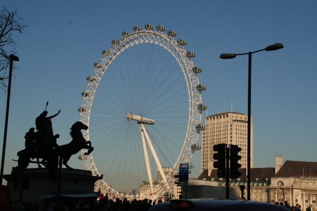 Ein großes Riesenrad befindet sich zentral im Bild, mit Gebäuden und Pfählen auf der rechten Seite und einer Skulptur auf der linken Seite.