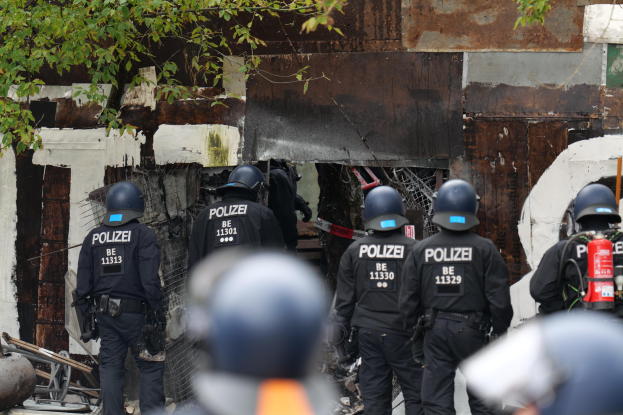 Eine Gruppe von Polizeiße in schwarzen Uniformen und Helmen steht vor einem Gebäude, mit einem Feuerlöscher auf der rechten Seite und Bäumen im Hintergrund.