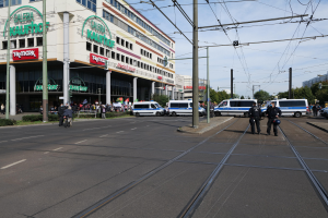 Polizeibeamte auf einer Straße neben einer Straßenbahn mit Fahrzeugen, Strommasten, Gebäuden, Bäumen und einem bewölkten Himmel im Hintergrund.