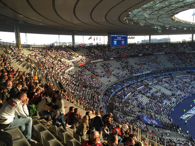 Eine große Menschenmenge sitzt im Allianz Arena in München, Deutschland, bei einem Fussballspiel, mit einer Bühne auf der rechten Seite, Fahnen, Stangen und einem Bildschirm im Hintergrund und dem Himmel oben sichtbar.