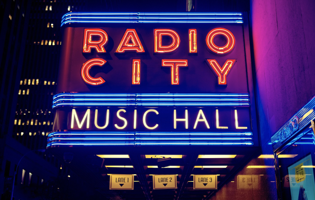 Außenansicht des Radio City Music Hall in New York City bei Nacht, hell erleuchtet mit hohen Gebäuden im Hintergrund, mit einem 'Radio City'-Schild und mehreren kleineren Schildern auf der rechten Seite.