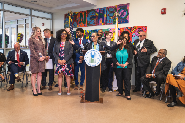 Eine Gruppe von Menschen, die vor einem Podium mit Mikrofon und einer Tafel mit der Aufschrift "Sancho City Councilman Joins City Council" stehen und sitzen, mit Flaggen, Feuerlöscher und Deckenleuchten im Hintergrund.