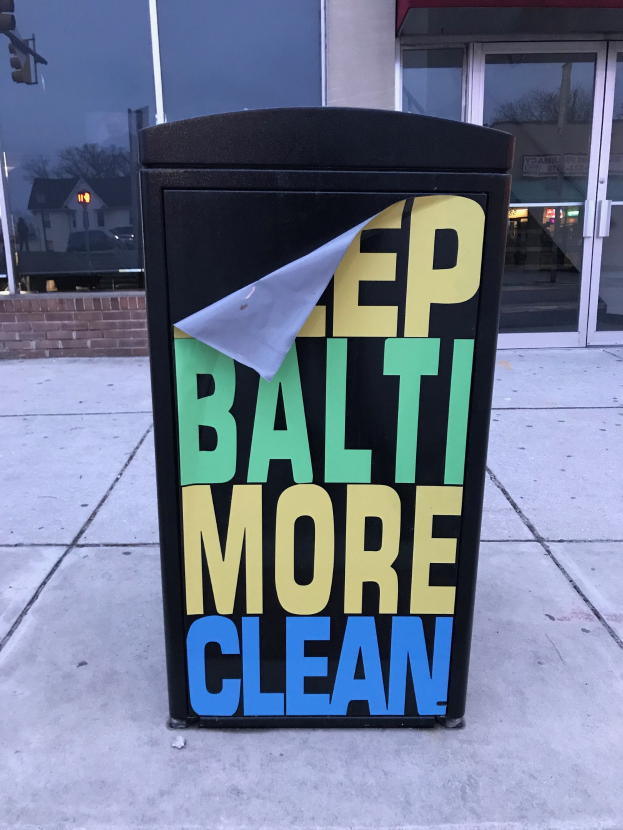 Schwarzer Müllcontainer mit "Keep Baltimore More Clean"-Text auf einem Gehweg vor einem Gebäude mit Glastüren.