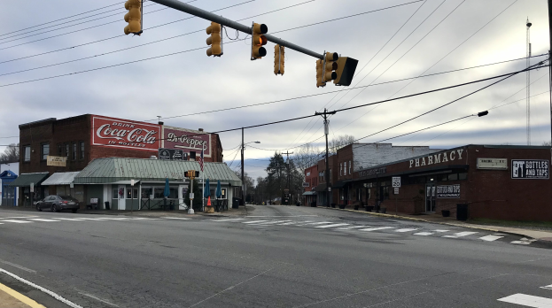 Stadtstraßenkreuzung mit einer Ampel, Fahrzeuge, Gebäude mit Beschriftung, Strommäste und -leitungen, Bäume und bewölkter Himmel.