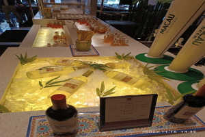 Ein Tisch mit Weinflaschen und Eiswürfeln, umgeben von Pflanzen und Brettern, bei einem festlichen Outdoor-Event mit Menschen, Tischen, Stühlen und Glastüren im Hintergrund.